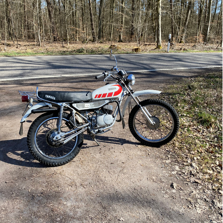 Eine klassische Yamaha Geländemaschine steht auf einem Waldweg, umgeben von Bäumen.