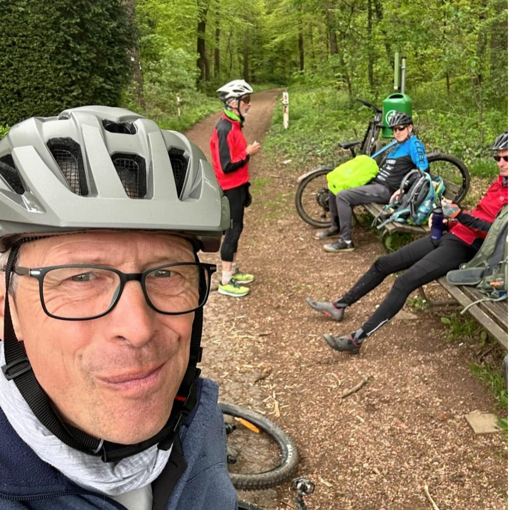 Vier Radfahrer machen eine Pause auf einem Waldweg. Ein Mann im Vordergrund macht ein Selfie, während zwei andere auf einer Bank sitzen und ein weiterer steht.