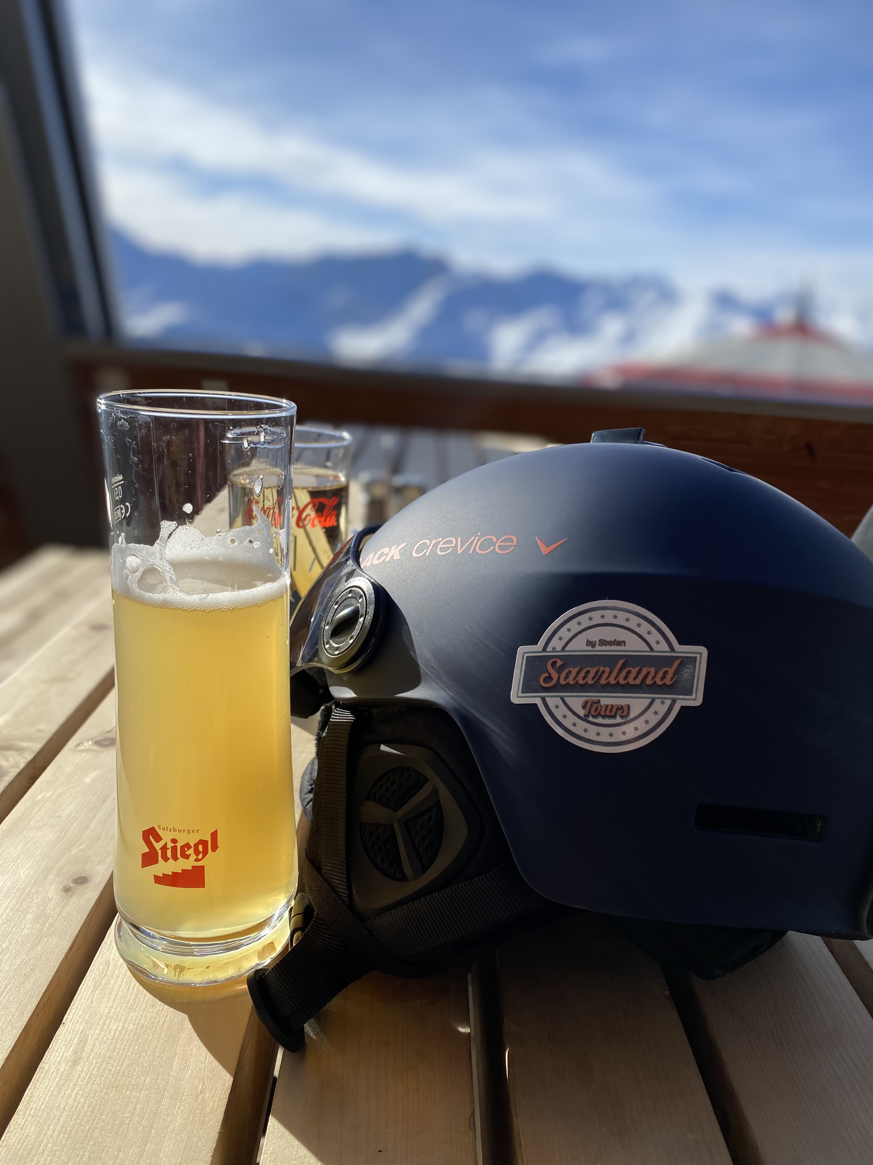 Ein Bierglas mit Stiegl-Logo steht neben einem Helm auf einem Holztisch mit verschneiten Bergen im Hintergrund.