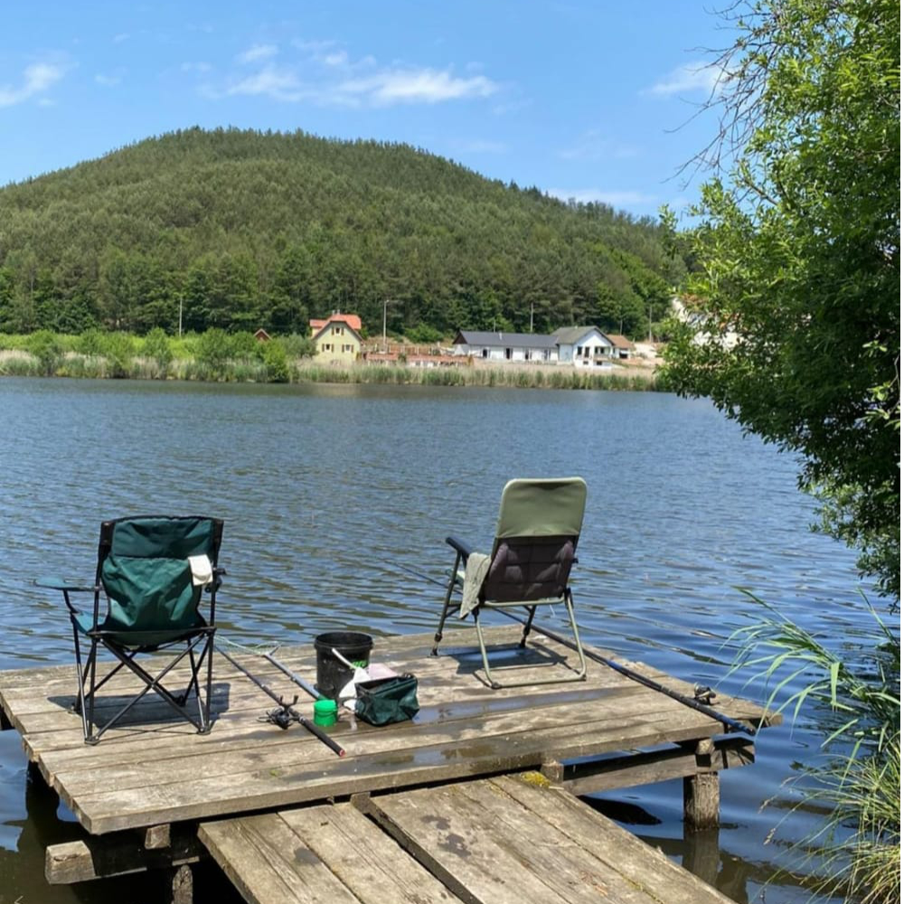 Zwei Anglerstühle stehen auf einem Holzsteg am See, umgeben von grünen Bäumen und einem Hügel im Hintergrund.