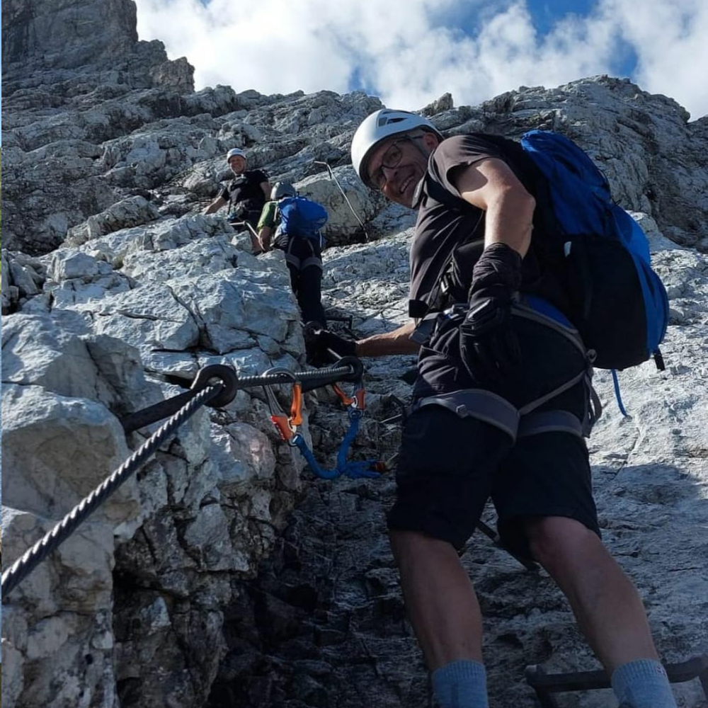 Kletterer erklimmen eine steile Felswand mit Kletterausrüstung und Helmen. Ein Mann im Vordergrund lächelt in die Kamera.